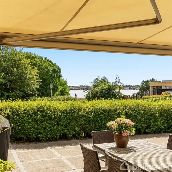 Terrasse med fliser og havemøbler. To liggestole står ved et med mursten opført hus med store vinduer. En høj, grøn hæk omgiver området. Solrigt skyfrit vejr.