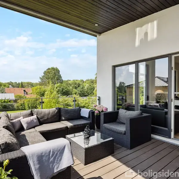 Terrassemøbler står på en overdækket altan. Møblerne er arrangeret omkring et lavt bord. Rummet åbner ud mod en frodig, grøn udsigt over nabolaget med blå himmel.