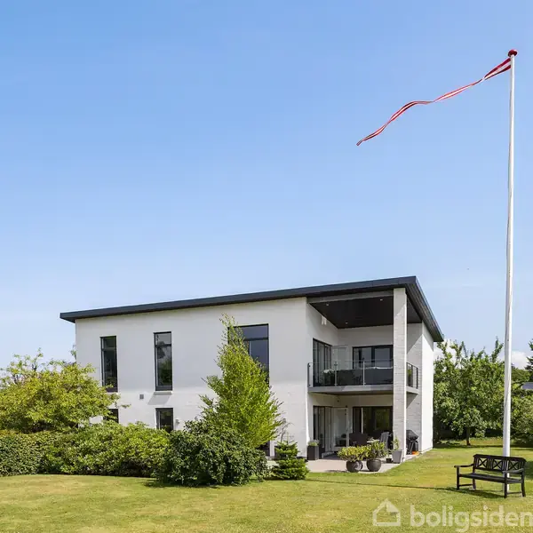 Et moderne to-etagers hus står på en velplejet græsplæne, omgivet af buske og træer. En høj flagstang med et flag ses til højre for huset. En bænk står ved flagstangen.
