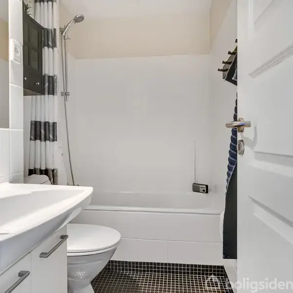 Håndvask med armatur ved siden af toilet, badekar med bruser i baggrunden. Badeværelset har hvide vægge, sort/hvidt forhæng og ternet gulvfliser. Dør står åben.