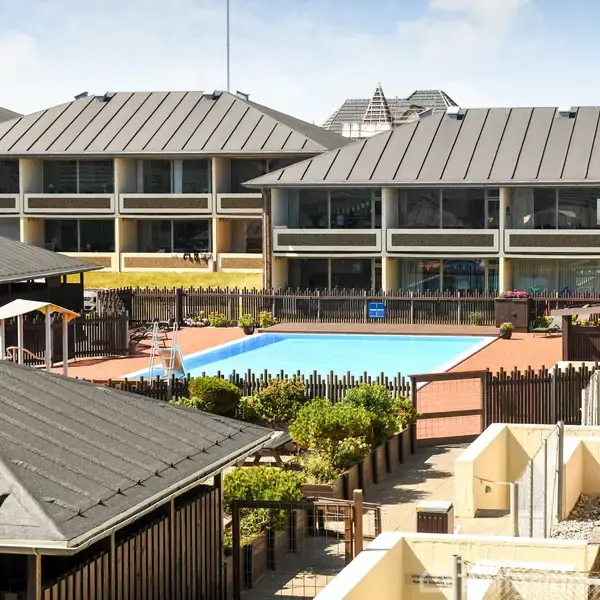 Beboelsesejendom med gråt tag omkranser en udendørs swimmingpool. Poolen er omgivet af hegn og beplantning. Baggrunden viser andre bygninger med skrå tage under en blå himmel.