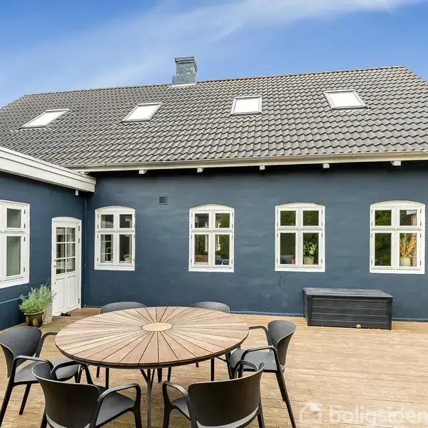 Et rundt træbord omgivet af sorte stole står på en træterrasse foran et blåmalet hus med hvide vinduer og en gråtag. Himlen er klar og blå.