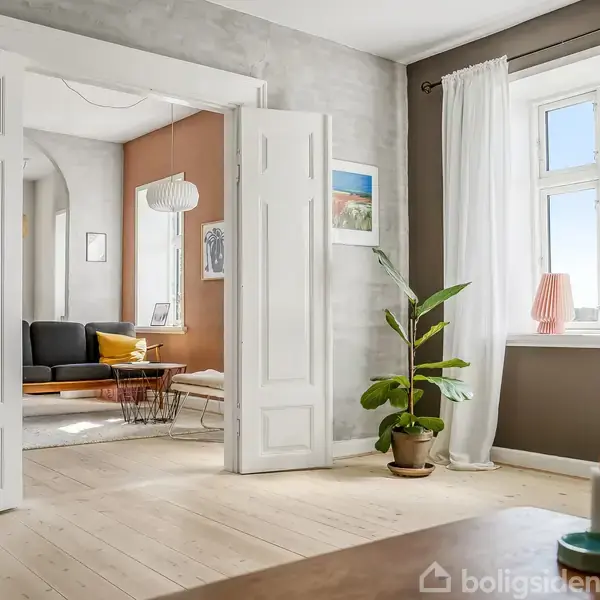 To åbne hvide døre leder til en stue med en mørk sofa og sofabord. Et stort vindue med gardiner slipper lys ind. En plante står ved siden af døren.