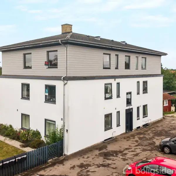 En hvid treetagers bygning med mørkegråt tag og vinduer. Den står i en gårdsplads med parkerede biler og omgivet af grønt.