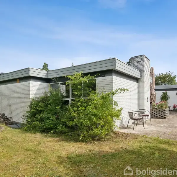 Et hvidt enfamiliehus står stille i en grøn have med en stenbelagt terrasse og to stole. Buske og træer omgiver huset under en blå himmel.