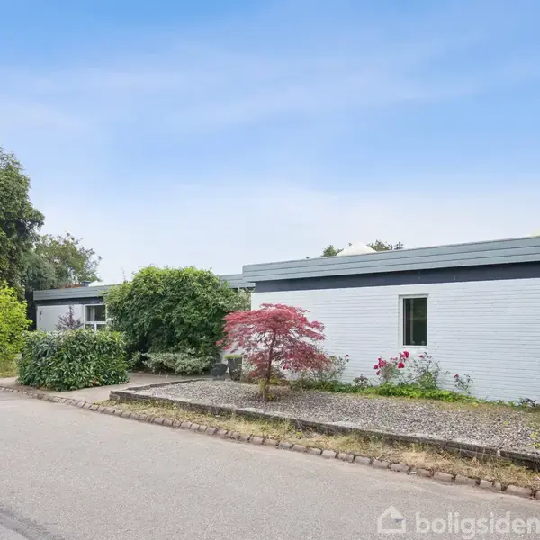 En hvid murstensbygning med fladt tag står stille omgivet af frodig vegetation og buske. Den ligger langs en stille vej med blå himmel i baggrunden.