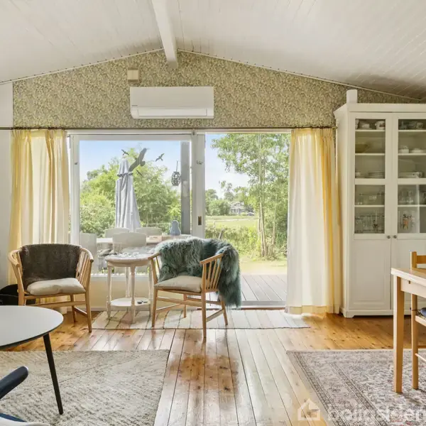 Stue med træmøbler og pejs. Store vinduer med gule gardiner åbner ud til en terrasse med parasol. Luftig indretning med tæpper på trægulv. Bright, enkel og moderne stemning.