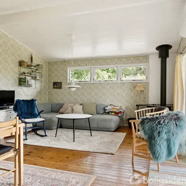 Stue med lyse møbler og trægulve. Sofa og sofabord står ved vinduerne. En brændeovn er i hjørnet, og spisebord med stole står til venstre. Overalt hænger kunst og hylder.