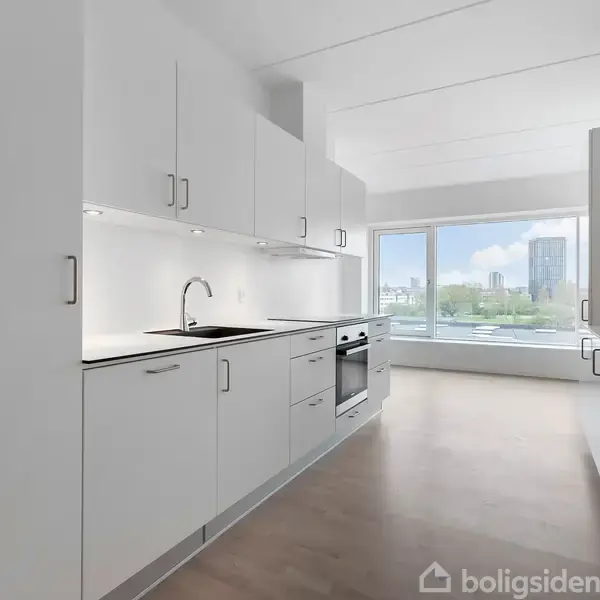 Et lyst køkken med hvide skabe og en vask. Rummet har trægulv og et stort vindue i baggrunden, der giver udsigt til en by med grønne områder.