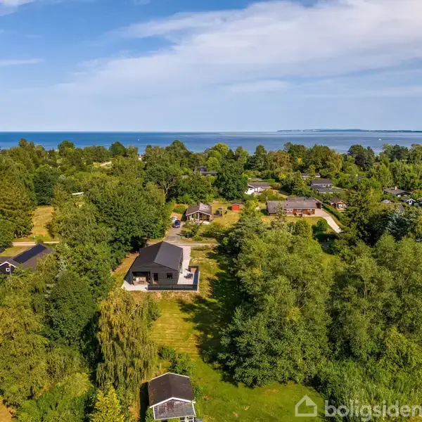 Hus omgivet af træer ligger i et grønt område nær kysten. I horisonten ses havet under en blå himmel. Der er flere huse spredt i det naturskønne landskab.