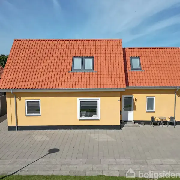 Et gult, to-etagers hus med rødt tag står på en flisebelagt gårdsplads. Sollyset skinner, og der er en grøn have ved siden af.