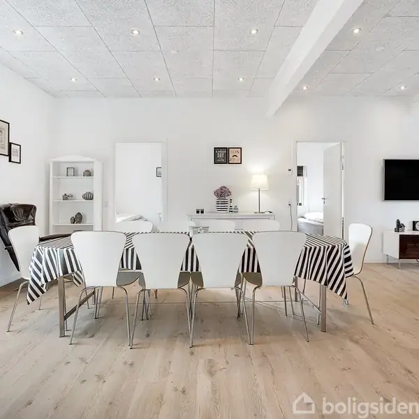 Et spisebord med hvidt mønsteret dug står i centrum, omgivet af hvide stole i en lys og moderne stue. Væggene har billeder, en reol, og der er en fladskærm.