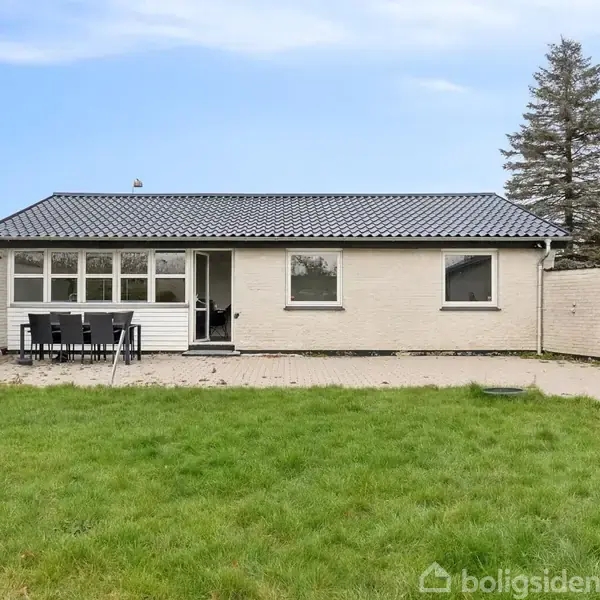 Et enkelt, fladt hus står på en frodig græsplæne. Der er et bord med stole på terrassen foran huset. Et højt nåletræ står til højre for bygningen.