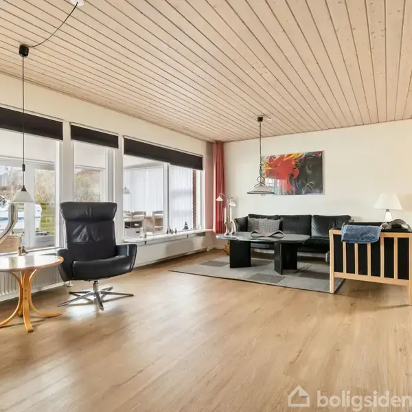 Stue med sort lædersofa og sofabord, flankerede af lænestole. Store vinduer i baggrunden giver udsigt til en terrasse. Træloft og gulv skaber en varm atmosfære. Maleri på væggen.