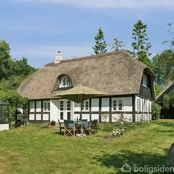 Stråtækt bindingsværkshus med hvid facade og sorte bjælker, står i en grøn have med træer omkring. Foran huset er et bord med stole under en parasol.