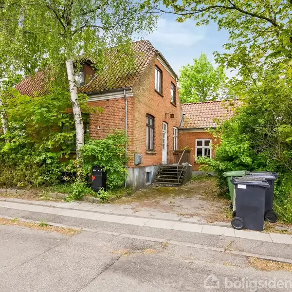 En rød murstensbygning med trappe foran, omgivet af grønne træer og buske. To skraldespande står på fortovet i en villavej.