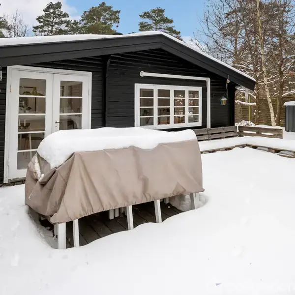 Et sort træhus ligger dækket af sne. Et bord med presenning står på terrassen, omgivet af sneklædte træer. En parasol står sammenfoldet til højre.