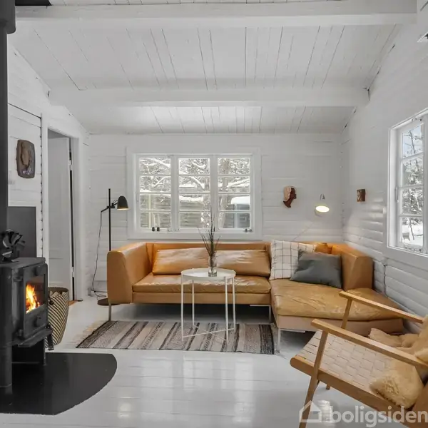Brændeovn står tændt, opvarmer et hyggeligt hvidmalet rum. En lædersofa og lænestol står på tæppe, mens sne ses gennem vinduet. En læselampe og lille bord fuldender miljøet.