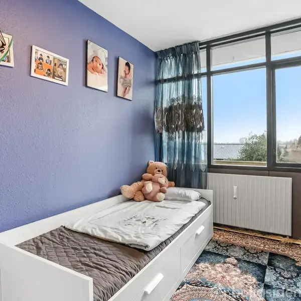 En hvid seng er placeret ved en blåmalet væg med billeder. En brun bamse sidder på sengen. Store vinduer giver udsigt til træer og himmel. Farverige tæpper dækker gulvet.