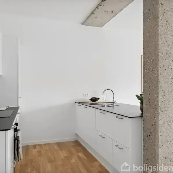 Køkken med hvidt interiør og sorte bordplader; plante, skærebræt og olier på bordet; betonbjælke som skillevæg; lyst trægulv. Spisestue med grå stole i baggrunden.