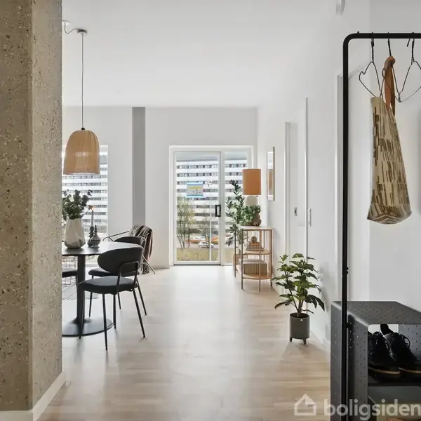 En moderne entré med en knagerække, plante og skohylde, der fører ind til en lys stue med spisebord, stole og pendel. Store vinduer viser en udsigt udenfor.