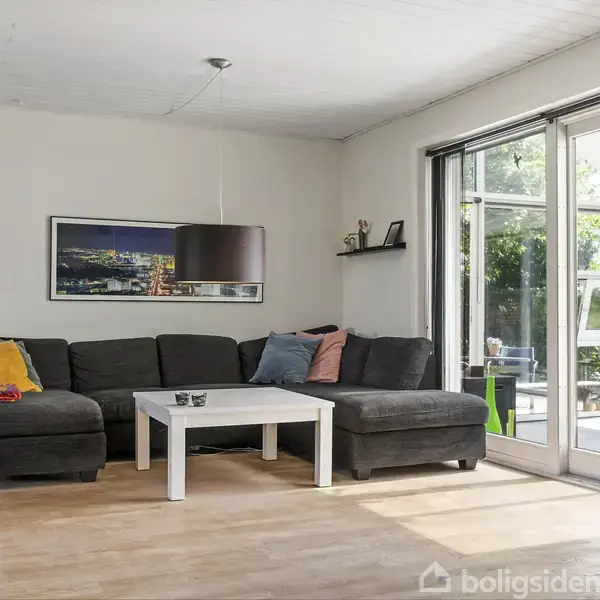 En grå hjørnesofa med farverige puder står i en stue ved siden af et hvidt sofabord. En stor skydedør fører ud til terrassen, hvor planter kan ses.