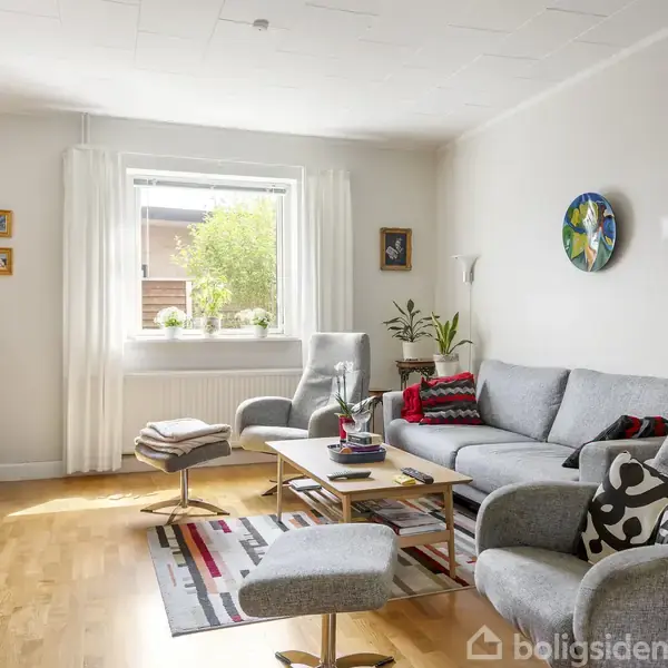 Stue med gråt sofamøblement placeret omkring et træbord med planter, omgivet af lyse vægge, billeder, og et fyldt reol. Store vinduer med udsigt til grønne planter udenfor.