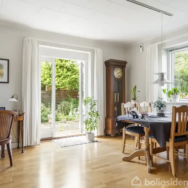 Et lyst spiseområde har et bord med stole, en høj skænk og en gammel standur. Gennem glasskydedøre ses en terrasse med grønne planter udenfor.
