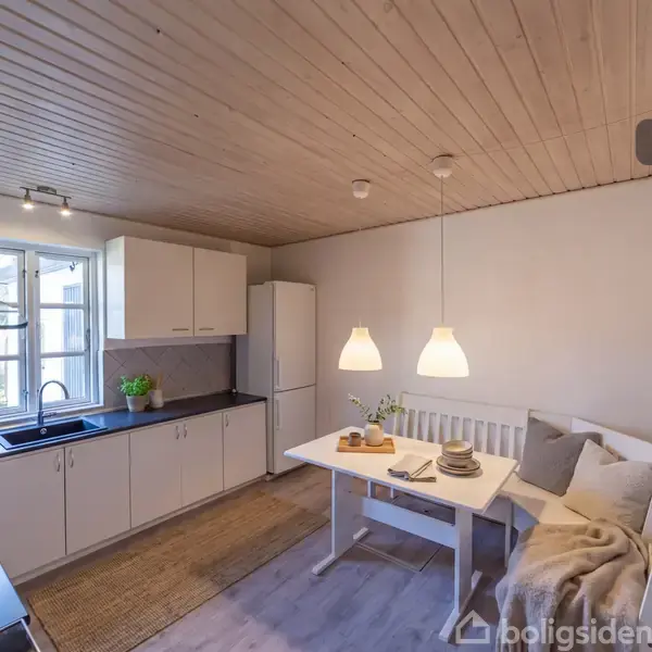 Et lyst køkken med hvide skabe og mørk bordplade. Et spisebord med to hængende lamper over står i rummet. Der er en bænk og stole ved bordet. Vinduet åbner mod haven. Tekst: "Visualisering".