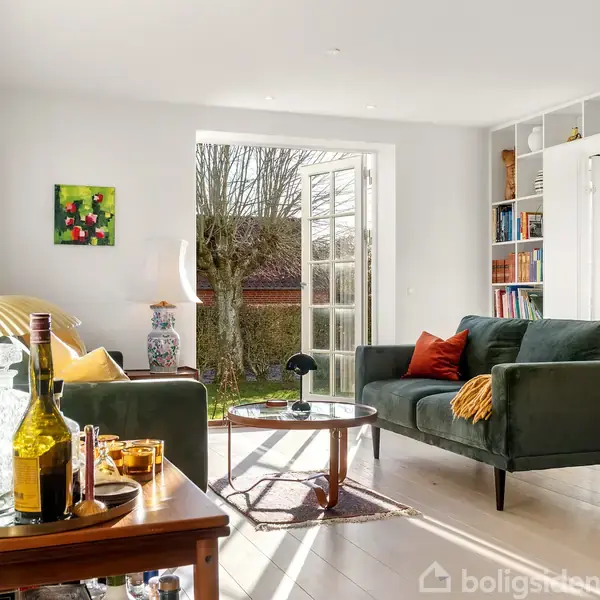 En stue med en grøn sofa og sofabord står ved en stor åbning til en have. Bogreol fylder højre side; et barbord med flasker er i forgrunden.