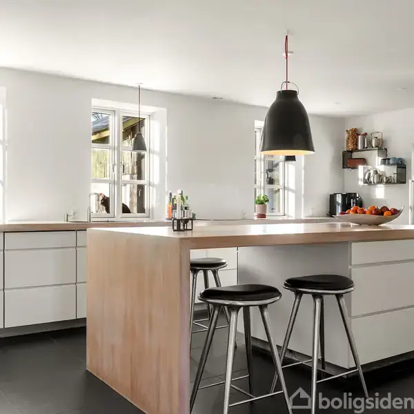 Et moderne køkken med en stor træø omgivet af tre barstole. Ophængte lamper hænger over øen. Store vinduer og hvide skabe udgør resten af miljøet.