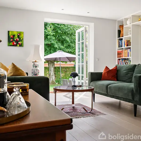Stue med grøn sofa, sofabord og reol fyldt med bøger; åbne franske døre fører ud til en have med parasol, hvor et maleri hænger på væggen, og en lampe står på et sidebord.