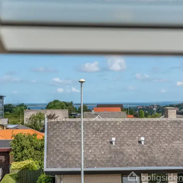 Vindue viser landskab; tage af huse og en havudsigt i horisonten. Himlen er blå med få skyer.
