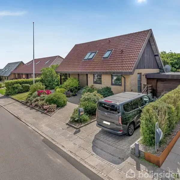 Et parcelhus står på en velplejet grund med en indkørsel, hvor en bil holder parkeret. Der er veltrimmede hække og blomsterbede, og det ligger i en rolig villavej.