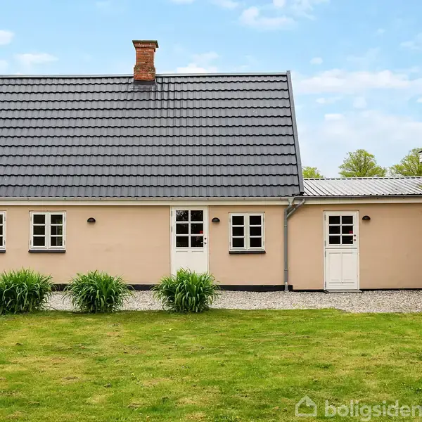 Et beige hus med sort tag står stille og roligt. Det har tre hvide døre og fire vinduer. Omgivet af en grøn plæne og blå himmel.