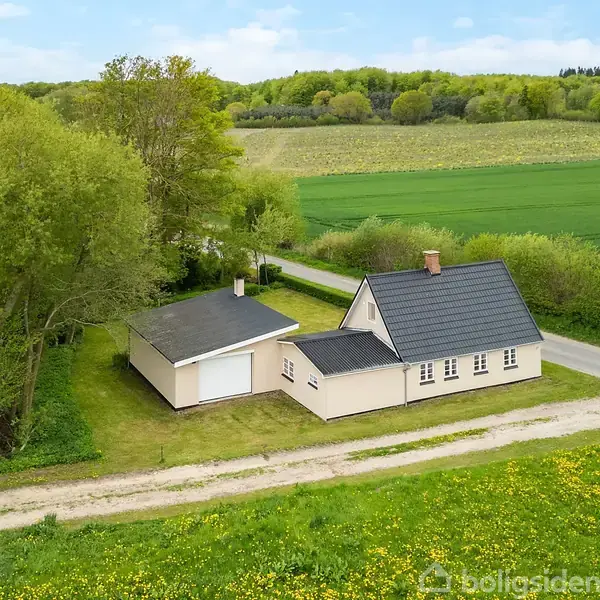Hus med mørkt tag står ved en grusvej, omgivet af grønne træer og marker i landligt område, der skaber en fredelig atmosfære.