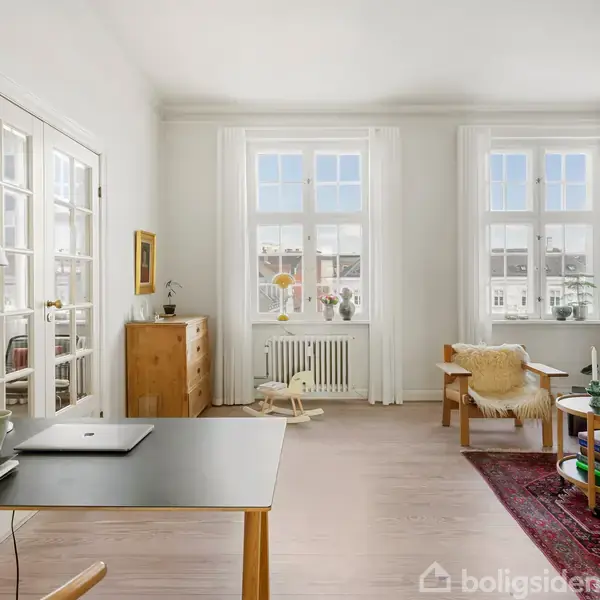 Et lyst rum med træbord og bærbar computer, kigger ud på to store vinduer. Sofa og stol står på tæppe ved siden af lille rundt bord. Radiator under vinduerne.