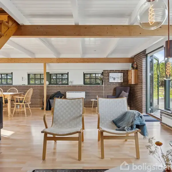 To træstole står i en stue med glasvægge. Til venstre ses en trappe, og i baggrunden er der et spisebord med stole. Lys strømmer ind udefra.