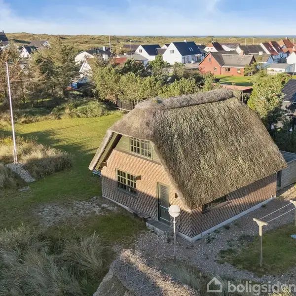 Et stråtækt hus står stille på en græsklædt grund omgivet af andre huse i et kuperet landskab med træer og buske.