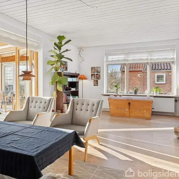 Et stueområde med to polstrede stole ved et bord dækket med sort dug. Omkring står planter, bøger på reol og lamper. Sollys skinner ind, og glasdøre fører til en veranda.