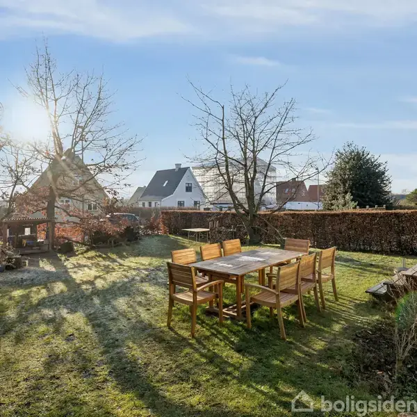 Et træbord med seks stole står på en græsplæne i en solrig have. Omkring er der træer og en hæk. I baggrunden ses huse og en blå himmel.