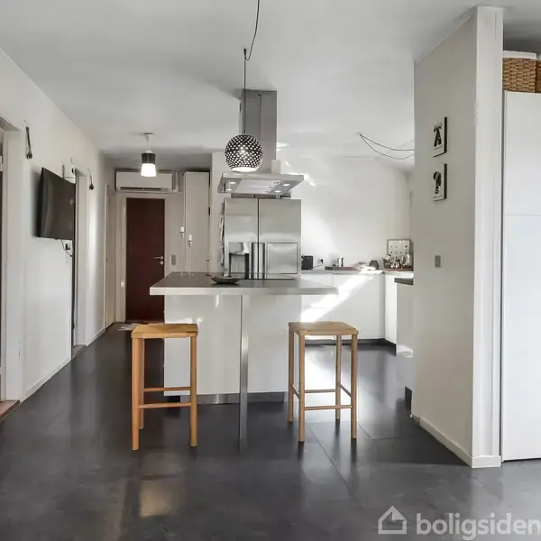Køkken med to høje barstole ved en central køkkenø. Køkkenet har hvidt inventar, køkkenhætte og lyse vægge. En åben gang er til venstre, med mørke gulve gennem hele rummet.