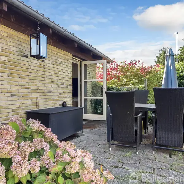 En terrasse med et bord og fire stole i kurvemøbler står foran et murstenshus. Omkring terrassen er der blomster og grøn beplantning, hvilket skaber en hyggelig atmosfære.