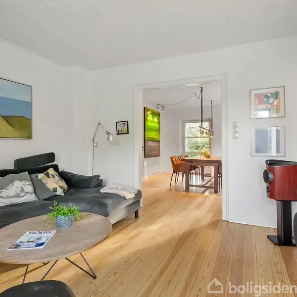 En stue med en sort sofa, sofabord, og plante på bordet. Stuen fører ind til en spisestue med et bord og stole. Vægkunst hænger omkring. Ingen tekst til stede.