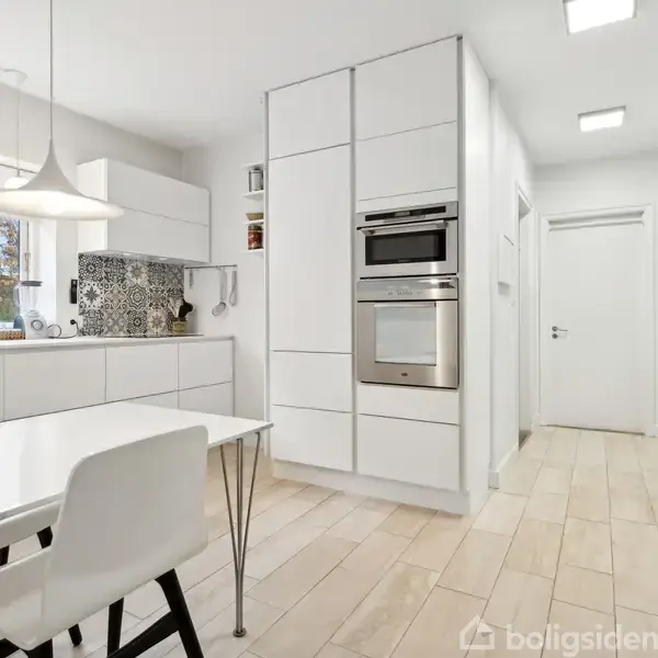 Køkken med hvide skabe og indbygget ovn; et hvidt spisebord med stole står i forgrunden. Store vinduer giver naturligt lys, mens loftslamper hænger over bordet. Fliser på gulvet.