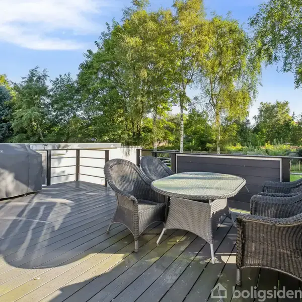 En terrasse med fire fletstole og et rundt bord står stille. Omkring er grønne træer og buske i en frodig have. Himmel er klar med få skyer.