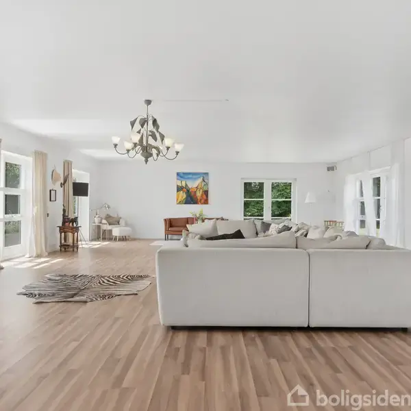 Stor, lys stue med hvide vægge og trægulv. Hvid sofa i midten, TV på venstre væg, lysekrone hængende fra loftet. Reol til højre. Store vinduer giver naturligt lys.