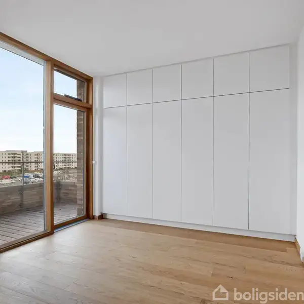 Dør fører ind i et tomt rum med trægulv, store indbyggede hvide skabe og et stort vindue mod en balkon og udsigt over andre bygninger.