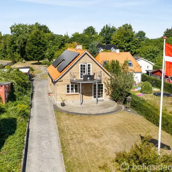 Et to-etagers hus med rødbrune tegl og sorte tagsten står på en velplejet græsplæne med en flagstang og dansk flag. Omgivet af grønne træer og hække.