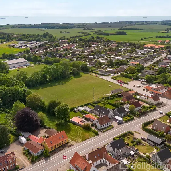 Luftfoto af en by med røde hustage, der omgiver et grønt område. I baggrunden ses marker og kystlinje. Bygader er synlige blandt beboelsesområderne.
